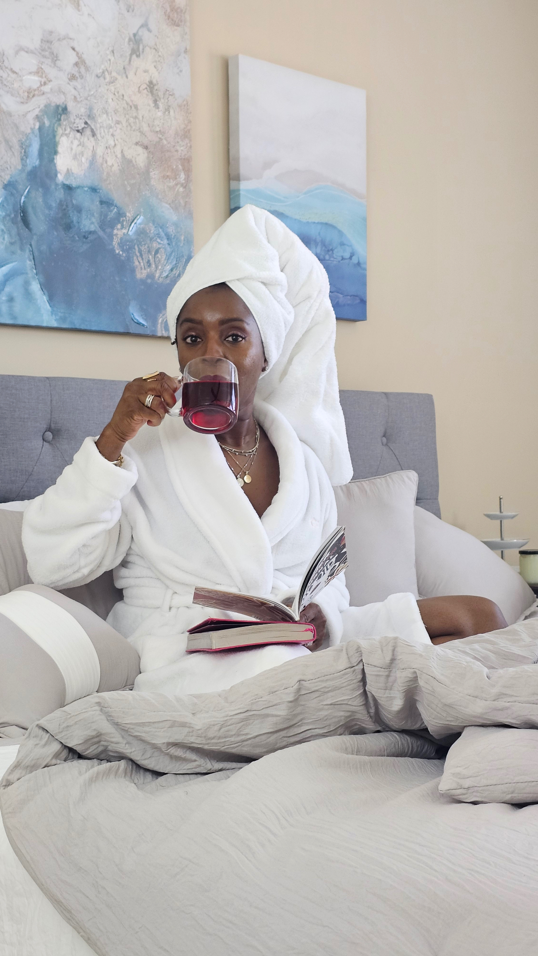 Mo James in a white robe and towel, sipping tea and reading a book in bed.